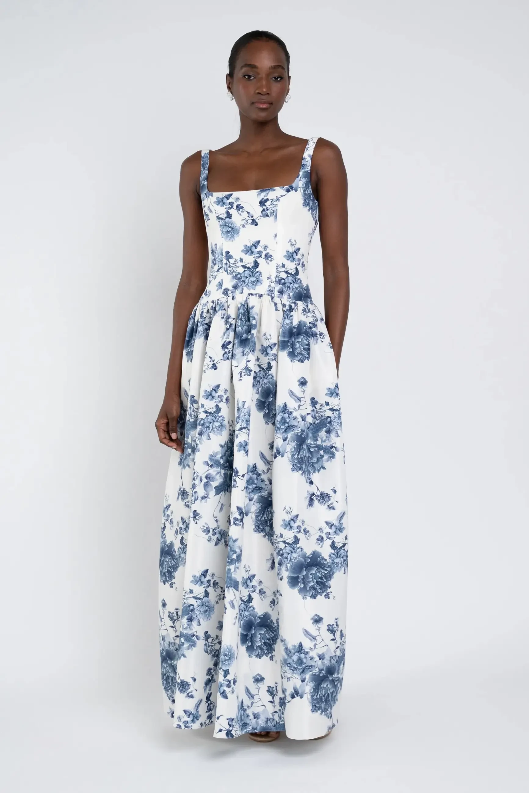 A woman in a white floor-length dress with blue floral patterns stands against a plain backdrop. The mood is elegant and serene.