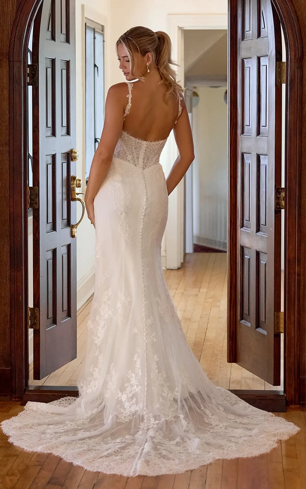 A woman in a white wedding dress standing by an open doorway, viewed from the back. The dress has delicate lace details and a train, and she has long hair styled in a ponytail. The interior features wooden doors and flooring.