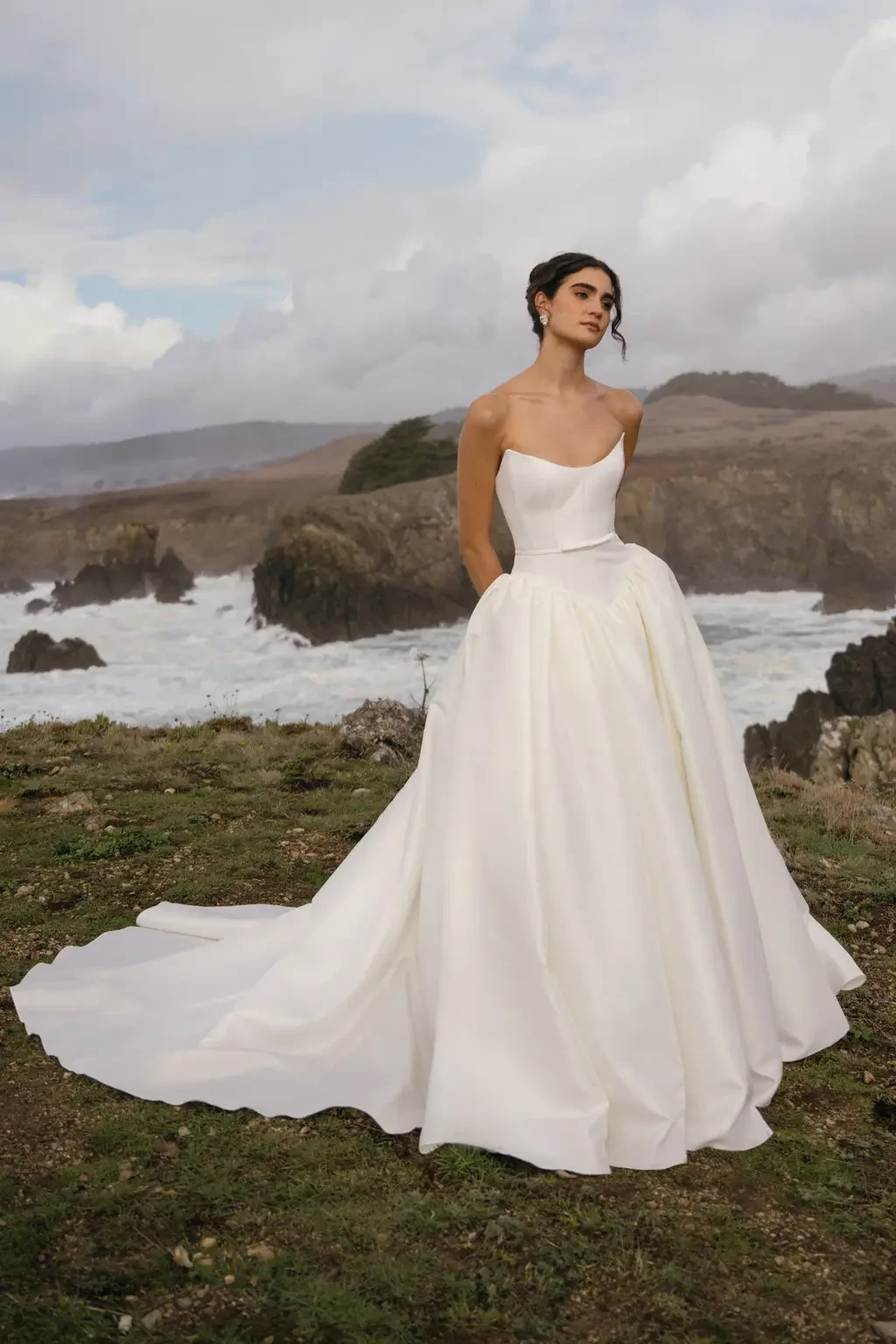 A woman in a strapless white wedding dress stands on a rocky landscape with the ocean and cloudy sky in the background.