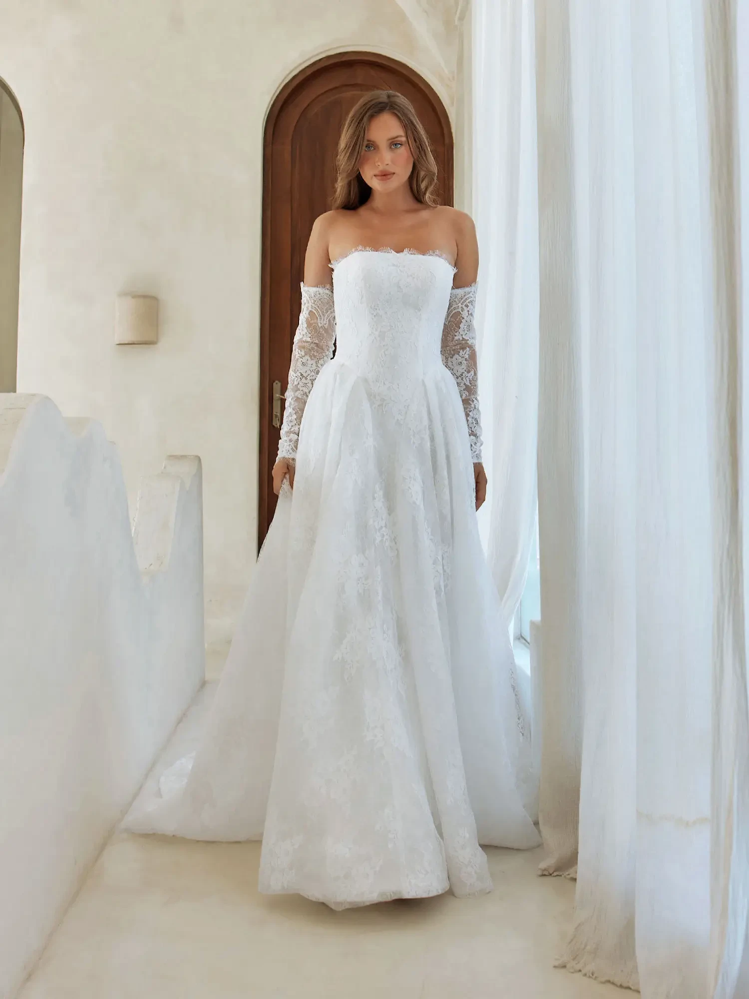 A woman wearing a white strapless wedding gown with off-the-shoulder lace sleeves, standing in a bright, airy interior with large windows.