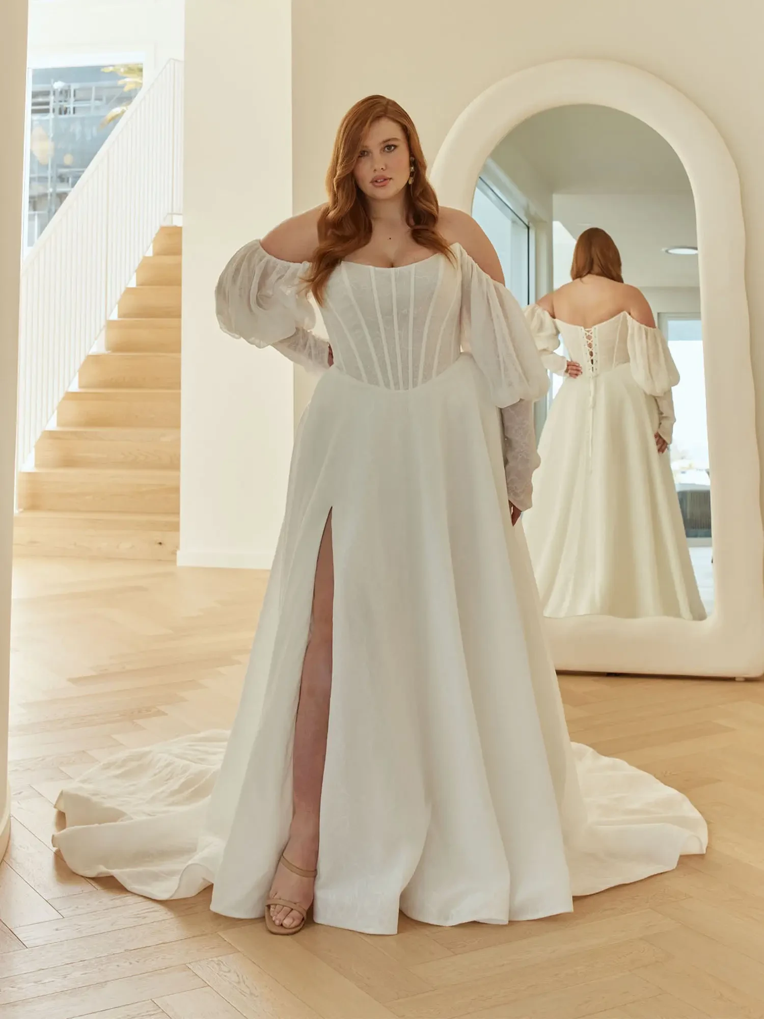 Woman in a white wedding gown with puffed sleeves and a slit stands confidently in a modern room, reflected in an arched mirror. Elegant, serene mood.