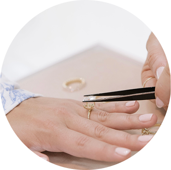 A person's hand is holding a pair of tweezers, delicately adjusting a ring on another hand. There are additional rings visible on the surface nearby.