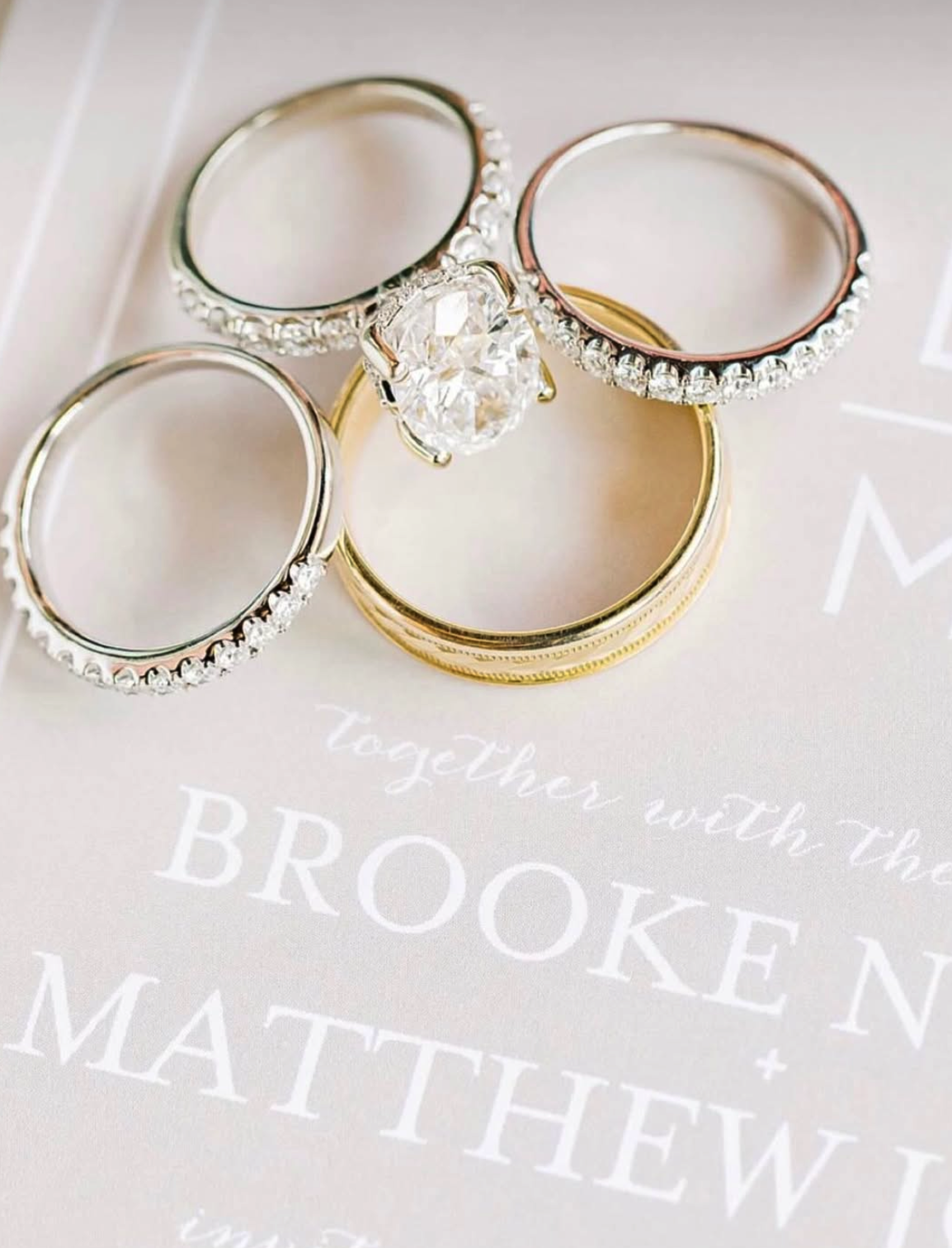 A close-up image of four rings arranged on a white surface with text. One ring features a large, prominent diamond surrounded by smaller stones, while the others are metal bands.