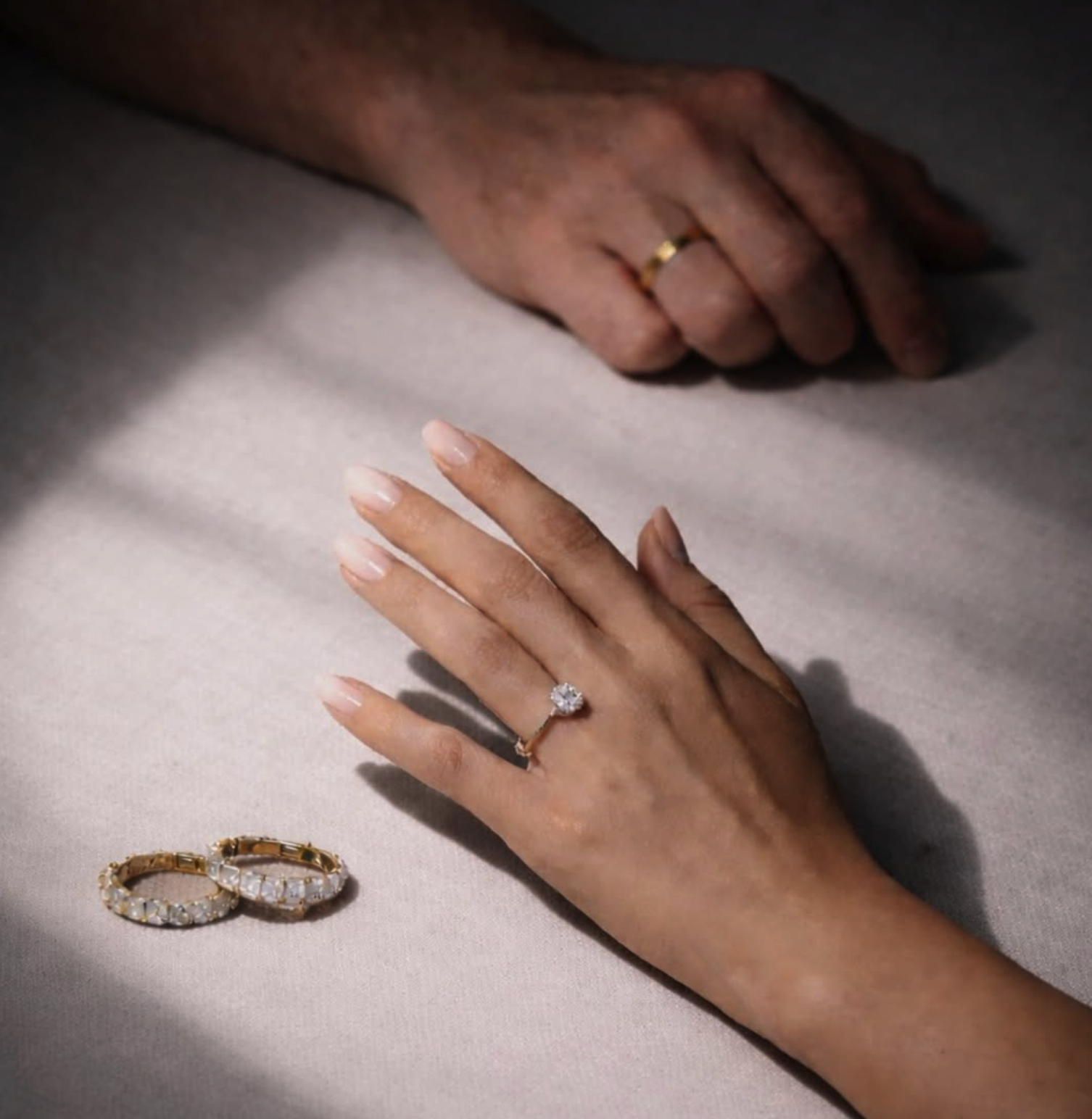 A close-up of two hands, one wearing a gold wedding band and the other showcasing an engagement ring with a large diamond. Two additional rings are placed on the surface nearby.