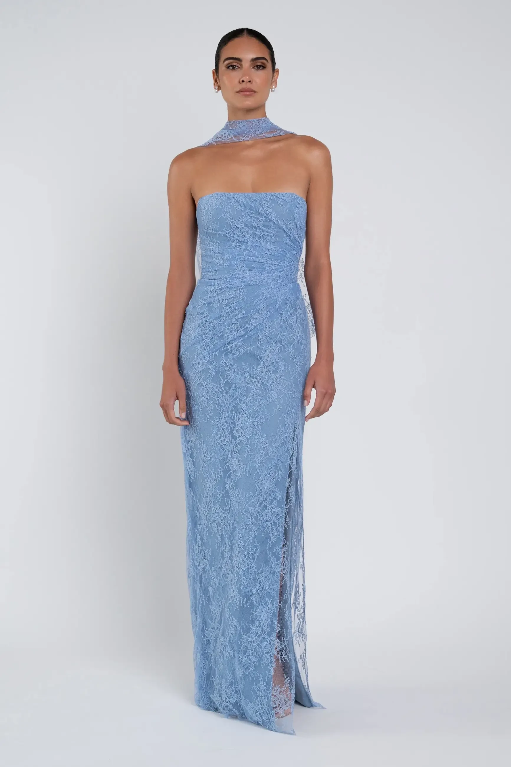 A woman in a strapless, blue lace gown poses against a plain, white background. The dress features a floor-length design with a side slit, and a matching neck wrap adds elegance.