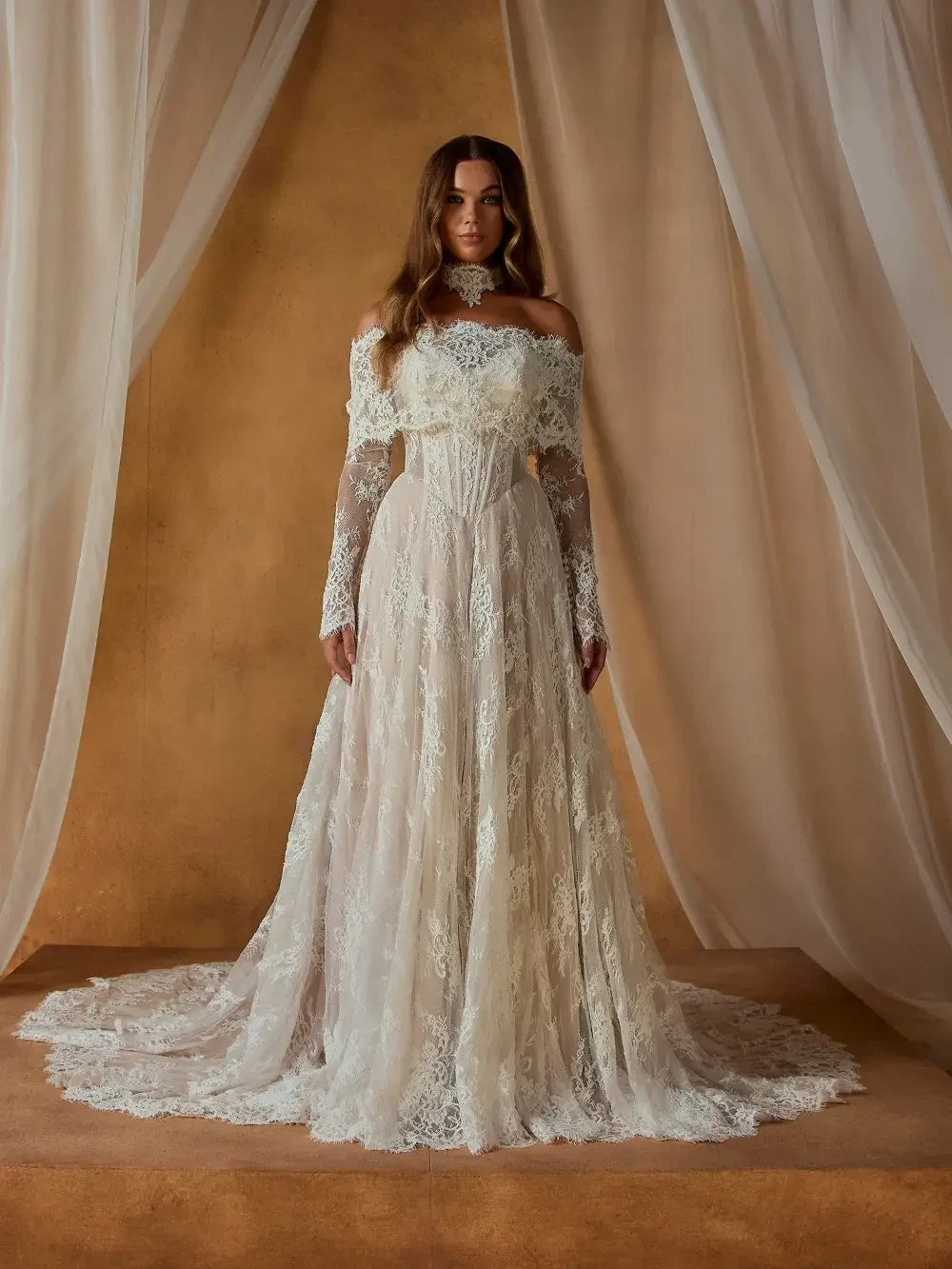 A woman in a strapless, lace wedding dress stands elegantly against a soft, neutral background.