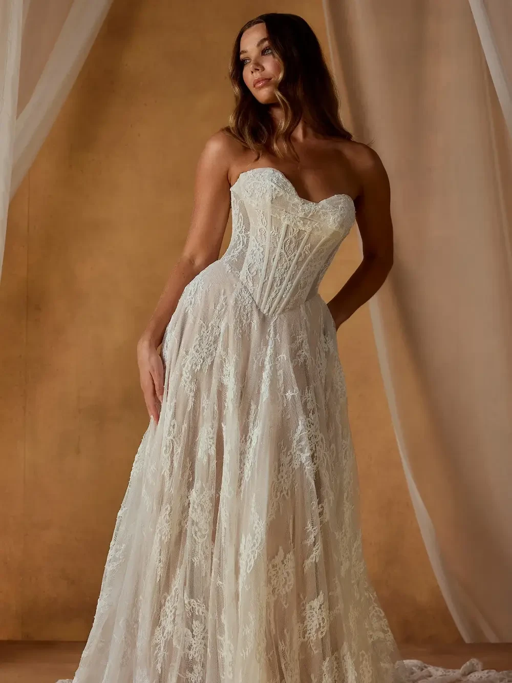 A woman wearing a long, elegant, off-the-shoulder lace wedding dress with long sleeves, standing against a warm-toned background with draped fabric.