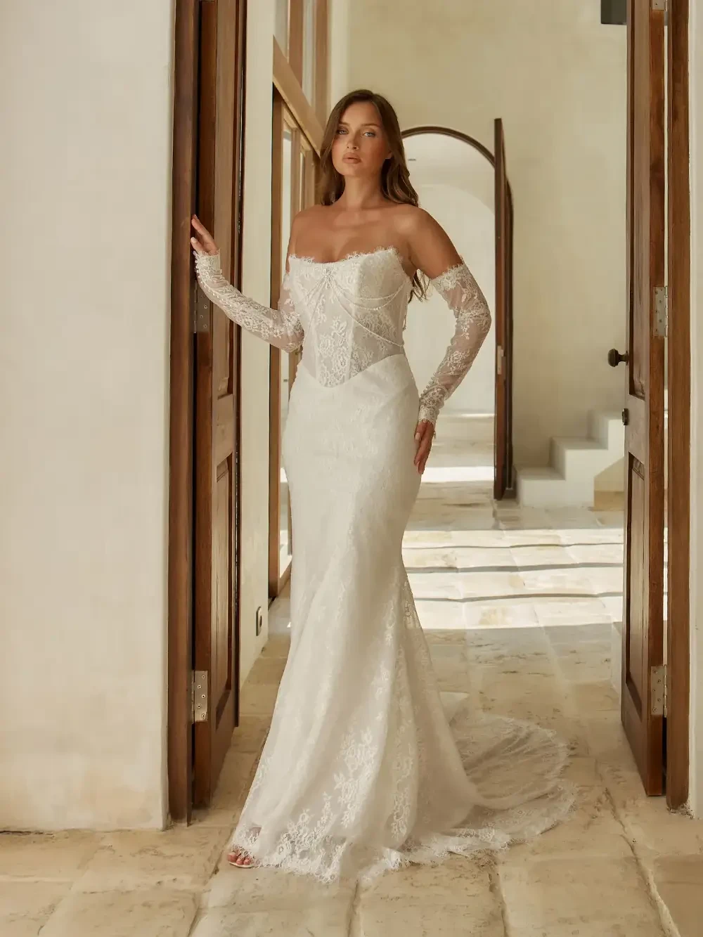 A woman in a white wedding dress with an off-the-shoulder design and lace detailing stands in a stylish interior with wooden doors.