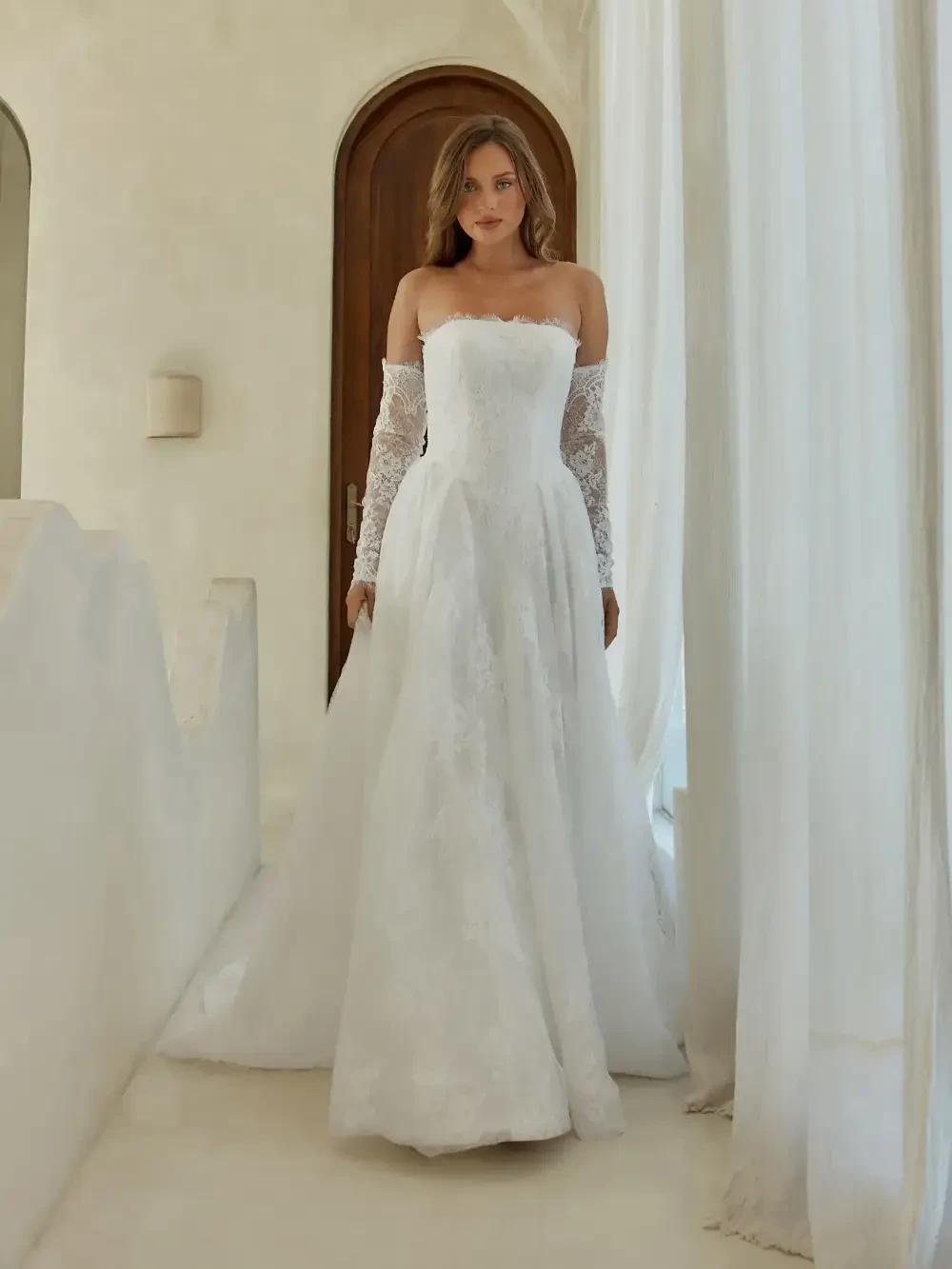 A woman wearing a strapless white dress with lace sleeves, standing in a well-lit room with white walls and a wooden door.